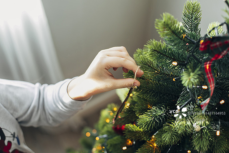男孩用装饰品和圣诞彩灯装饰圣诞树。寒假快乐的男孩在家里装饰圣诞树。圣诞节的家庭传统。图片素材