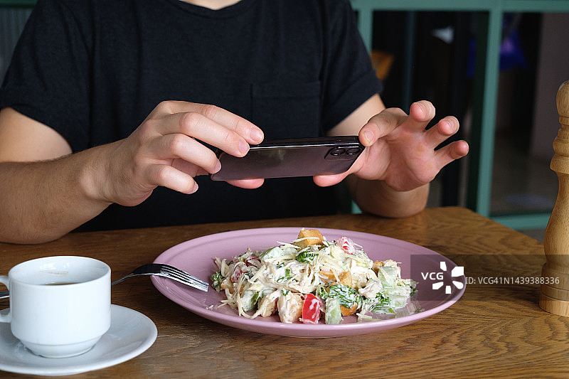希腊的沙拉。健康饮食的概念。沙拉放在盘子里，放在咖啡馆或餐馆的桌子上。一个男人在餐桌上吃沙拉，在家里喝咖啡。一名博主用手机拍下食物，然后用视频拍摄故事。图片素材