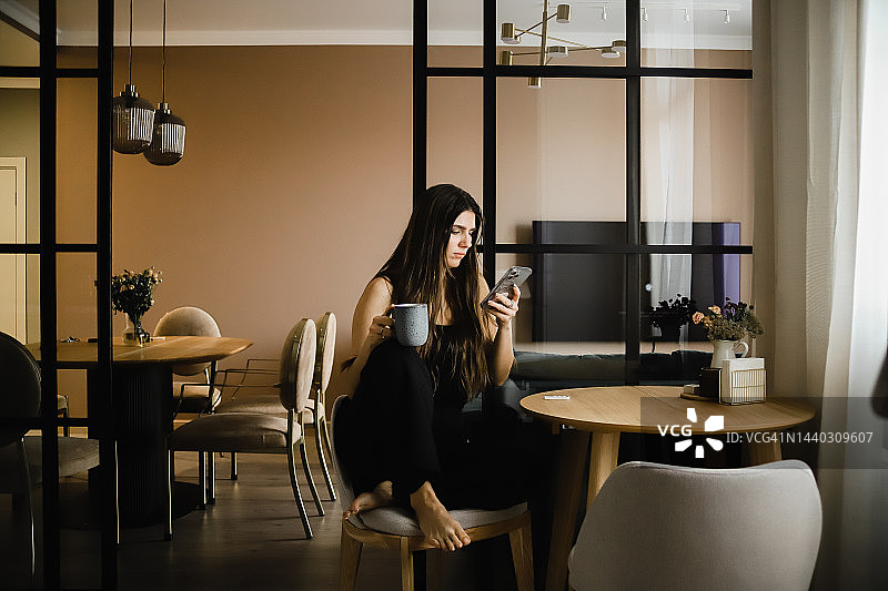 一个头发飘逸、穿着深色衣服的女孩坐在厨房里，用马克杯喝着热饮图片素材