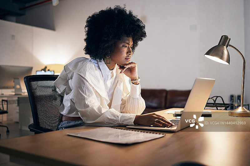 电子邮件，晚上和黑人妇女在笔记本电脑上，工作和阅读互联网上的建议在办公室工作。年轻的非洲企业员工在黑暗中用电脑制定战略图片素材