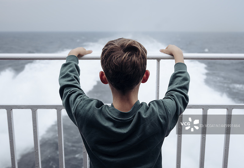 一个男生站在一艘渡船的栏杆上，看着轮船引擎喷出的水花图片素材