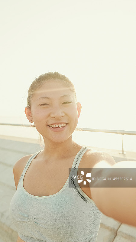 微笑的亚洲女孩在运动上衣自拍，站在堤岸在晨光图片素材
