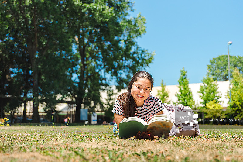 在一个阳光明媚的课后，微笑的女学生躺在草地上看书。世界读书日。阅读的好处图片素材