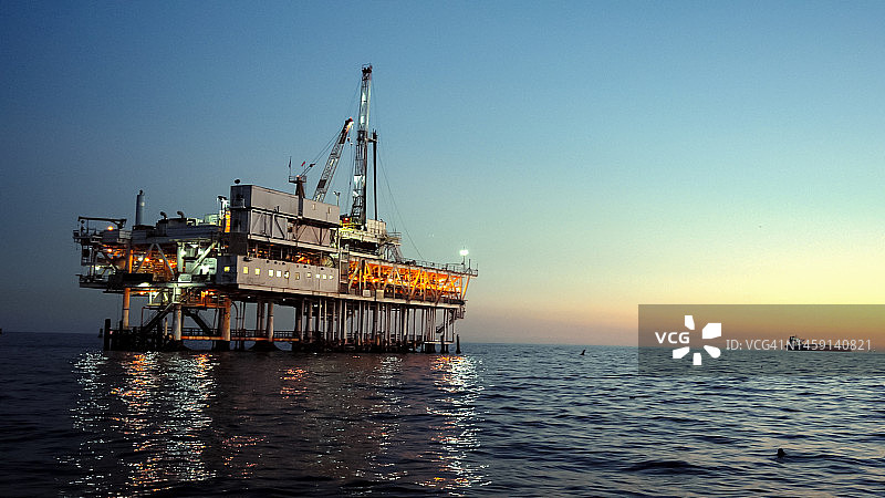 灯火通明的大型工业天然气和石油海上钻井平台在平静的海面上，在一个晴朗的傍晚，远处的背景是卡特琳娜岛图片素材