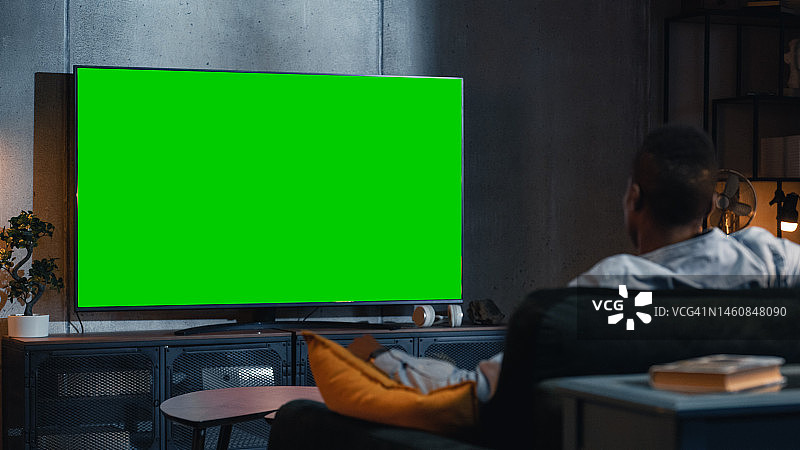 黑人坐在家里的沙发上，用Chromakey观看绿屏模拟电视显示器。放松的男性观看晚间电影。从肩膀上看。现代在线娱乐概念。图片素材