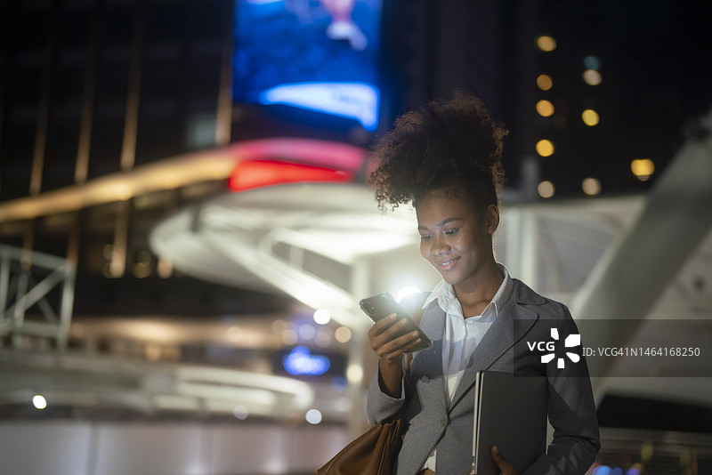 美丽的年轻女子使用智能手机走过夜晚的城市街道图片素材