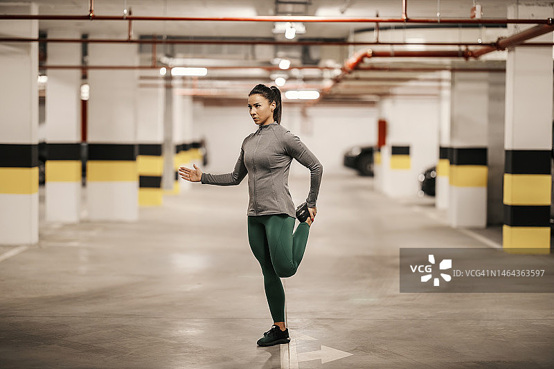 一位肌肉发达的女运动员在地下车库中单脚站立并伸展身体。图片素材