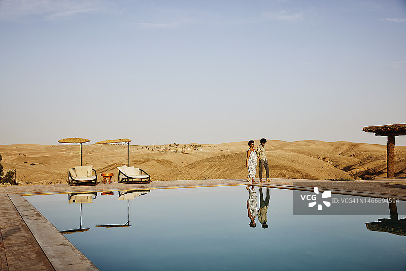 宽镜头的情侣散步在游泳池在沙漠营地日出图片素材
