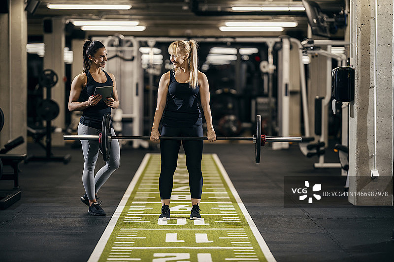一位身材健美的女健身运动员正在健身房举重，一位女私人教练正在看着她。图片素材