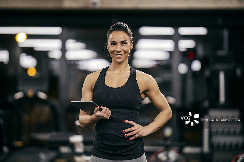 在健身房里，一位身材健美、肌肉发达的女健身教练正拿着平板电脑对着镜头微笑。图片素材