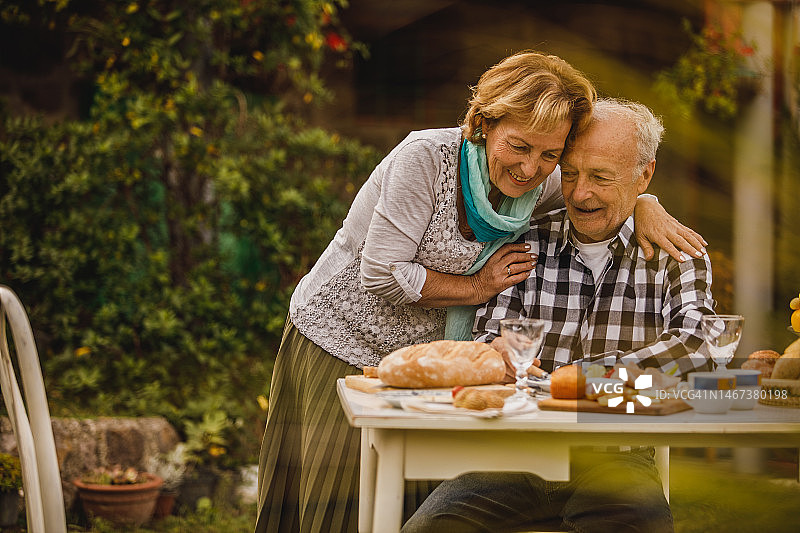 在后院吃晚饭时，一位富有爱心的老年妇女从后面拥抱着她爱的丈夫图片素材