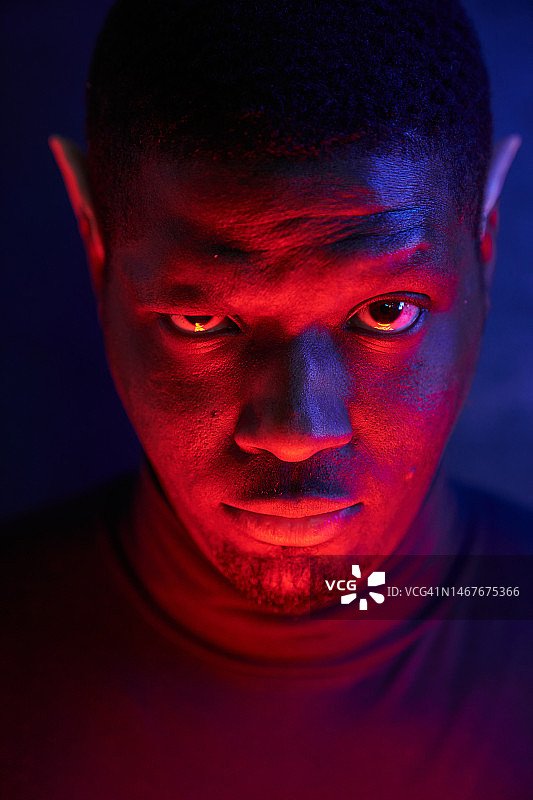 一个穿蓝红相间衣服，戴着精灵耳朵，打着霓虹灯的黑人。一个人的艺术肖像在一个黑暗的背景与复制空间。图片素材