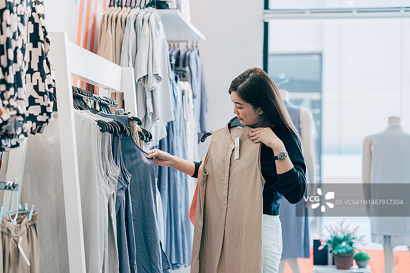 亚洲妇女试穿衣服做决定。一名女子在服装店购物时看着休闲服装。图片素材