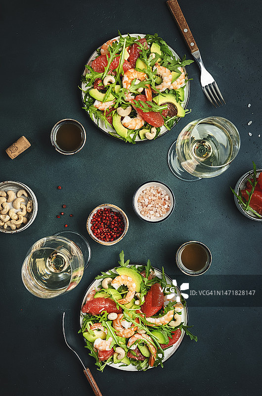 海鲜沙拉配虾仁、鳄梨、葡萄柚、芝麻菜和腰果。餐桌上摆放着白葡萄酒。深绿色背景，俯视图图片素材