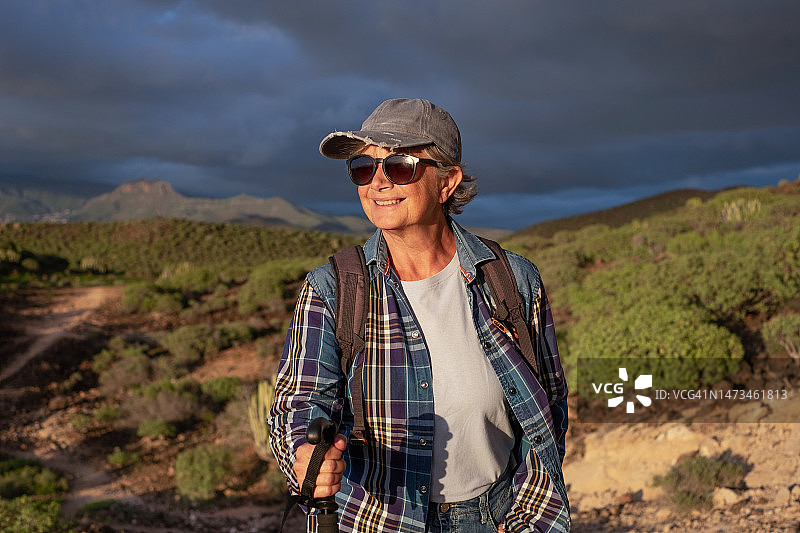 美丽的老年妇女在帽子和运动服在徒步旅行的日子走在日落享受自由和假期。快乐的成熟女性用棍子看着山地热带景观图片素材
