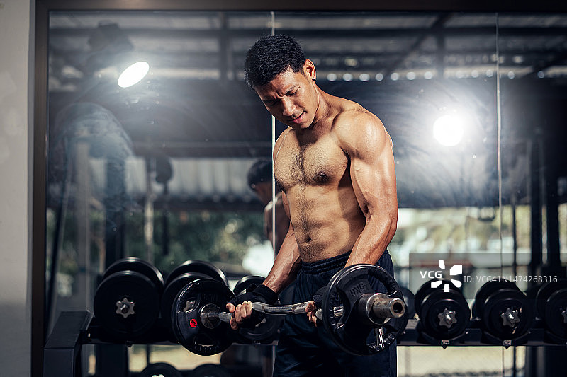 亚洲男子健美运动员赤膊努力重量训练肱二头肌卷曲与杠铃在健身房。图片素材