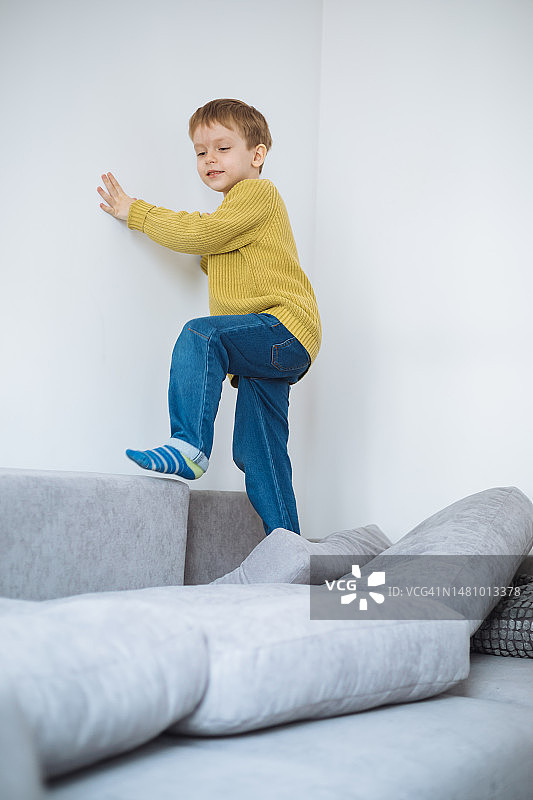 顽皮的金发男孩爬过灰色沙发上的靠垫，靠在白色的墙上图片素材