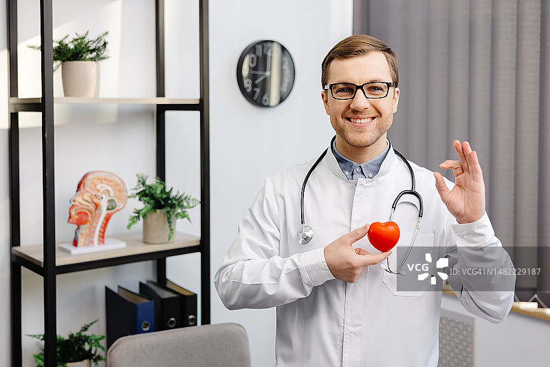医生穿着白大褂，戴着眼镜，手里拿着一颗红心站在医院办公室。医疗保健和医生员工的服务理念图片素材