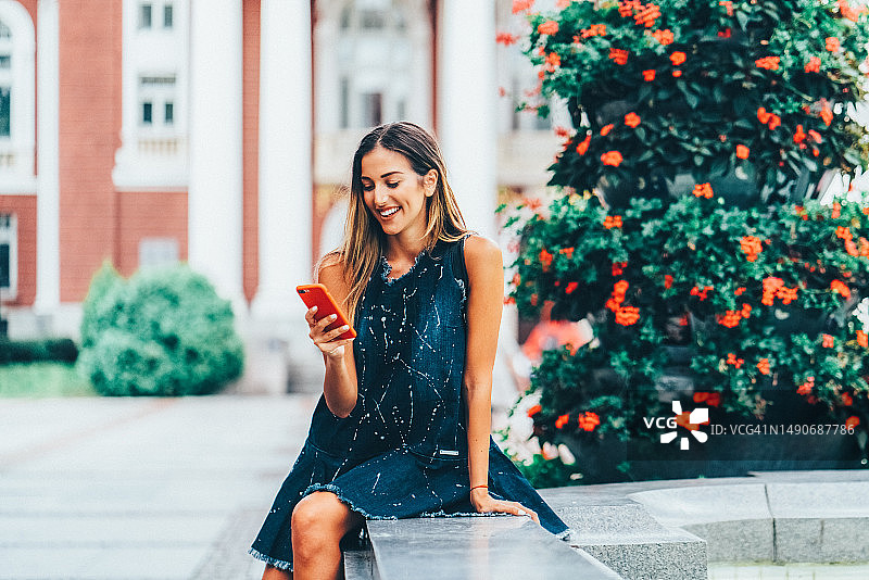 在炎热的夏天，一位微笑的女子坐在大学附近使用智能手机图片素材