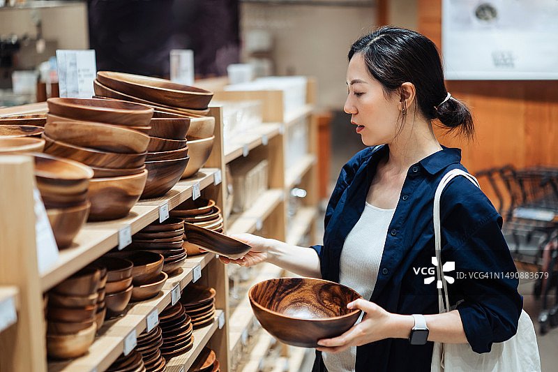 一名年轻的亚洲妇女在一家家居用品商店购买家居用品和生活必需品，看着货架上的木碗。零售及购物概念图片素材