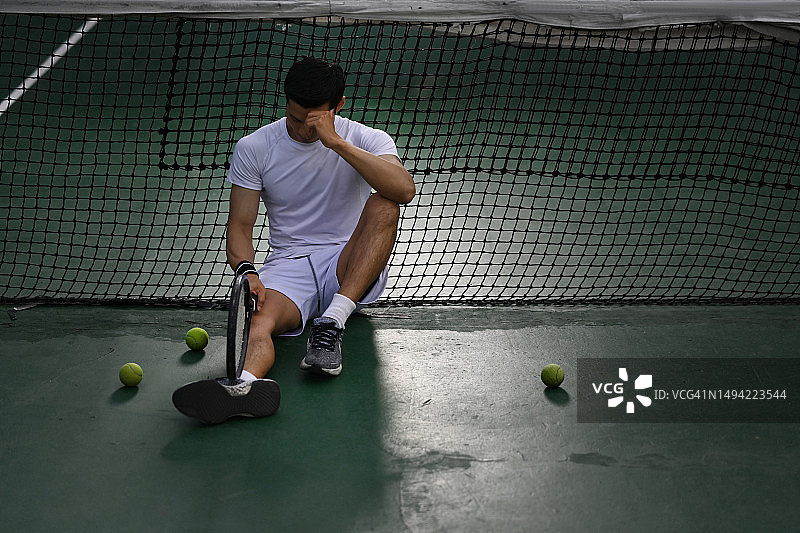 失望的亚洲男子网球运动员坐在球场上，心烦意乱的比赛失利，失败的情绪。图片素材