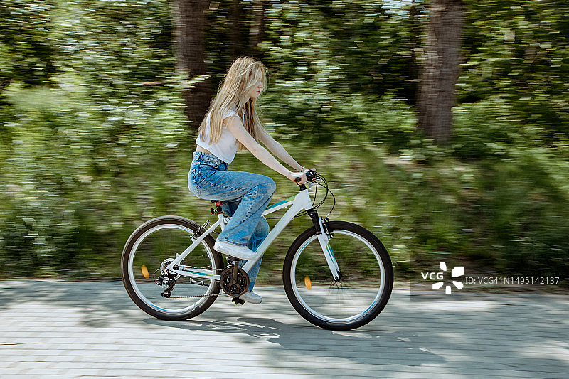 十几岁的金发女孩骑着白色的自行车在人行道上经过公共汽车和树木图片素材
