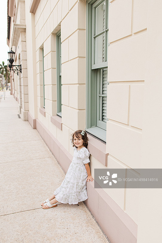 2023年5月，在佛罗里达州棕榈滩的一个托斯卡纳风格的花园中，一个4岁的古巴裔美国女孩，棕色卷发，穿着鼠尾草绿和白色的春季连衣裙，扎着发结，穿着白色的皮凉鞋图片素材