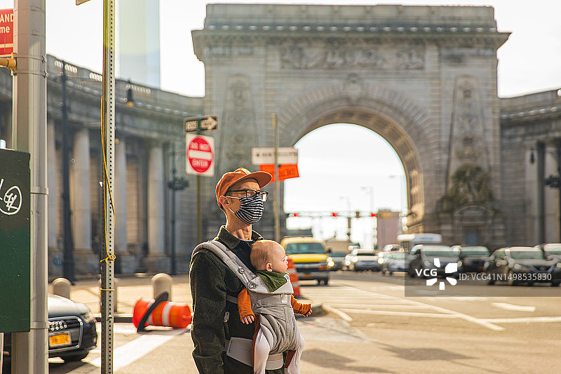 新冠肺炎期间，一名男子戴着口罩，用婴儿背带抱着女儿站在城市街道上图片素材