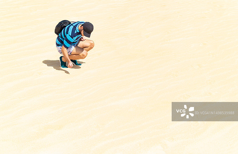 在一个阳光明媚的热天，一个年轻人蹲在沙漠的沙丘上图片素材