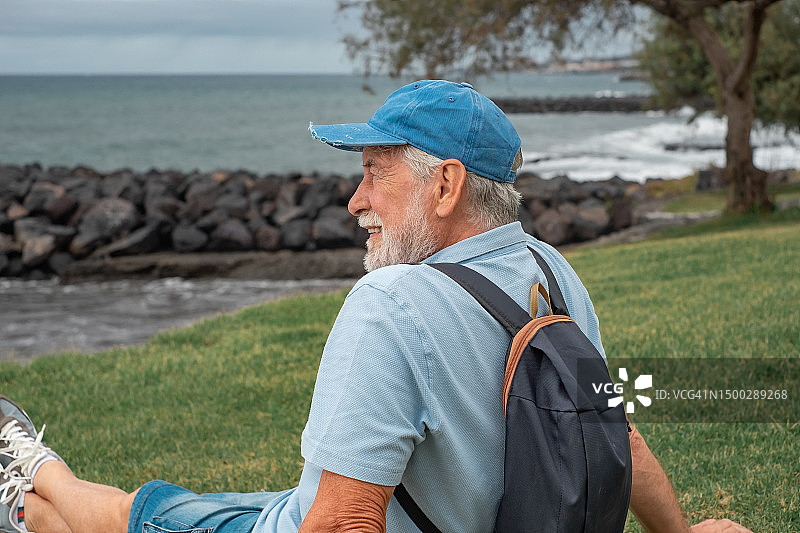 冬日里，微笑的老人坐在沙滩上的草地上，享受着休假或退休的自由时光。蓄着胡子、戴着蓝帽子的人看着海上的地平线图片素材