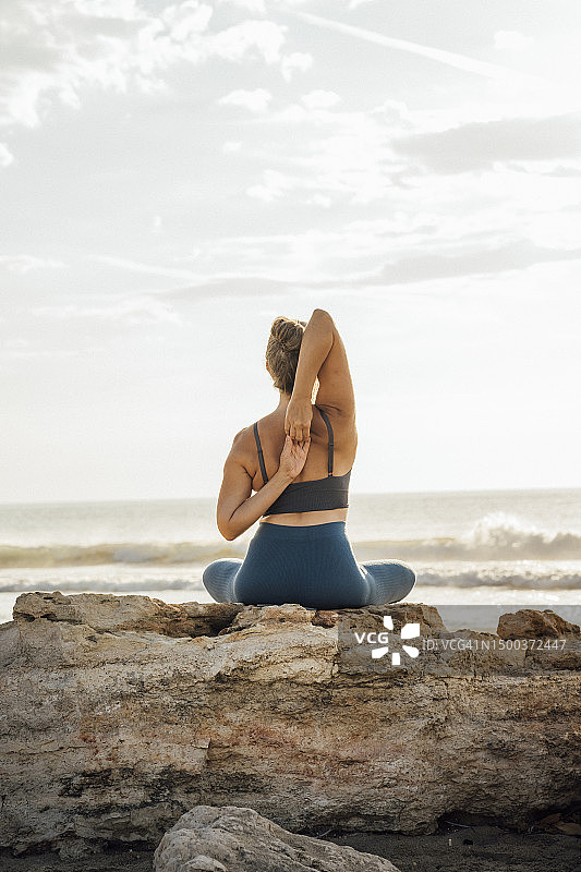一名女子坐在沙滩上的岩石上做瑜伽，双手放在背后图片素材