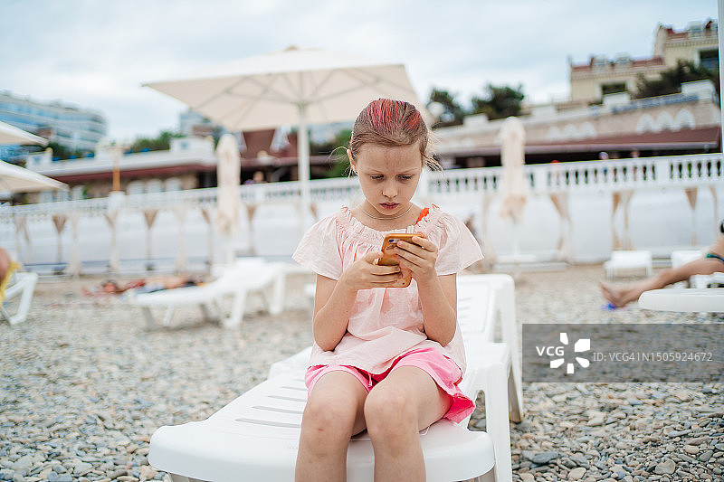 孩子坐在沙滩上的躺椅上玩智能手机图片素材