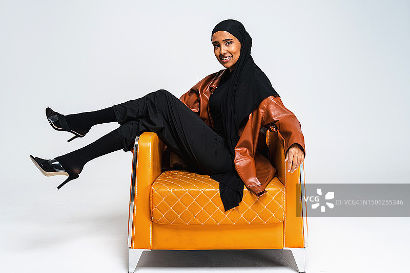 美丽的年轻女子穿着传统的中东服饰abaya坐在白色背景的工作室黄色沙发上图片素材