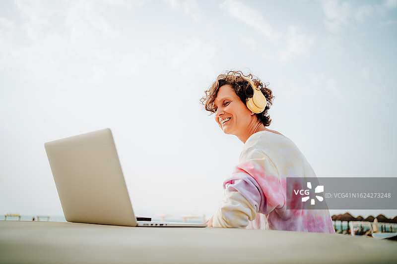 早晨的海滩娱乐无线连接:微笑的短发女人在她的笔记本电脑上视频通话，听音乐，同时沐浴在海滩上的阳光图片素材