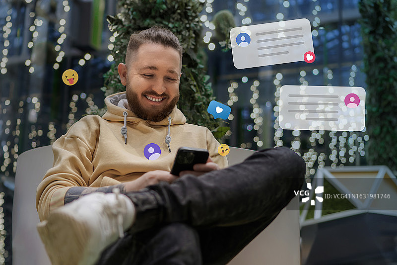 一个穿着米色卫衣、蓄着胡子的快乐男人的肖像，他正在用智能手机与朋友和同事交流。带表情符号的生活方式照片图片素材
