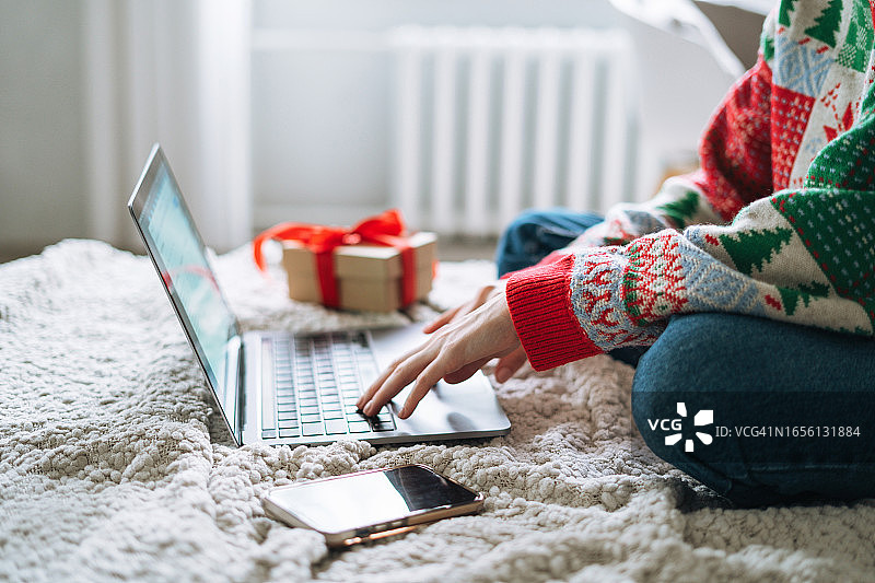 身穿红色休闲毛衣的年轻女子坐在家里的床上使用笔记本电脑，在网上圣诞购物图片素材