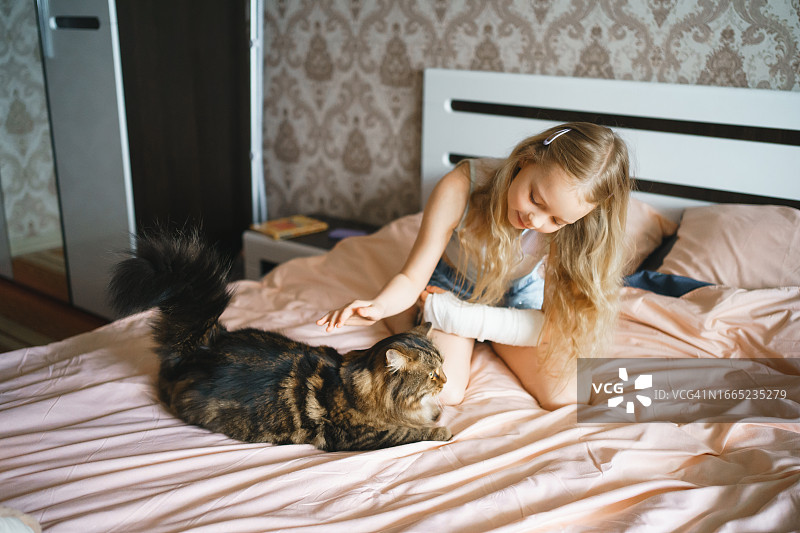 手臂上打了石膏的女孩在床上和一只猫玩耍图片素材