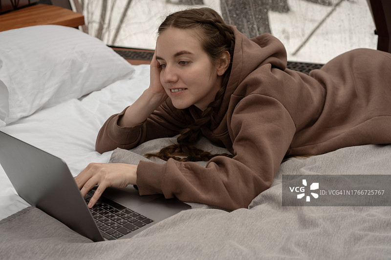梳着两条小辫的年轻女子正在上网，用笔记本电脑和智能手机工作。以冬季森林为背景的大型全景窗户。在家工作，现代科技图片素材