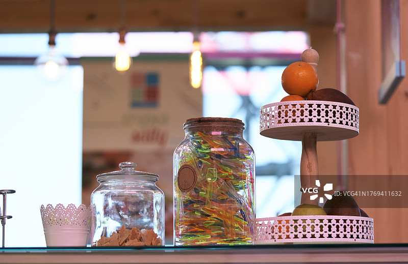 柜台上摆放着几个大小形状各异的容器，背景中有糖果、塑料棒棒糖和水果。图片素材