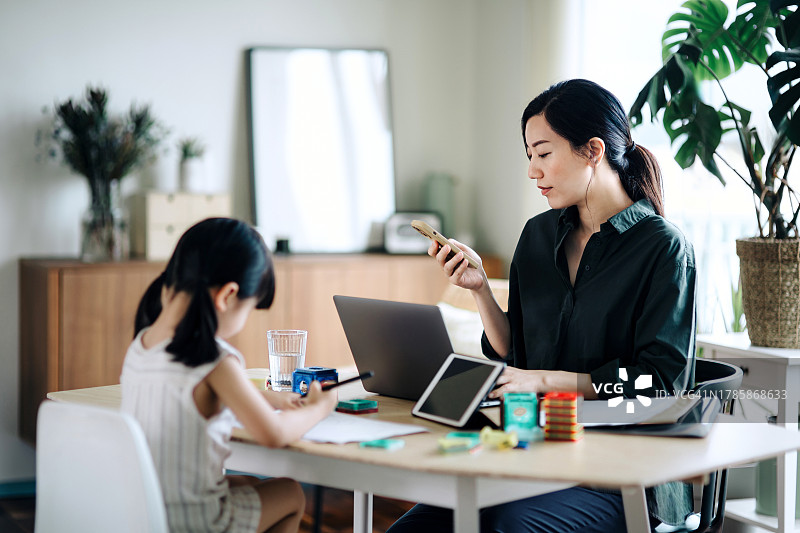 年轻的亚洲母亲在家里用笔记本电脑和智能手机工作，而小女儿在客厅学习。工作的母亲。母亲带着孩子在家工作。管理好工作和家庭生活的职业父母图片素材
