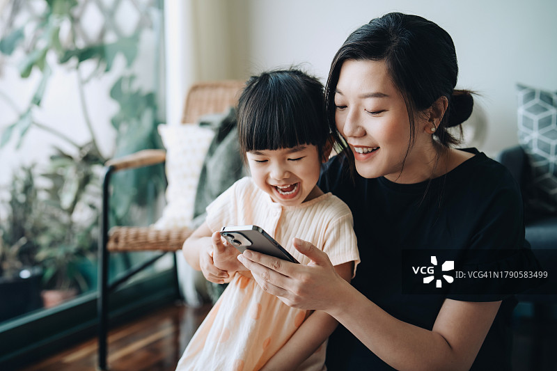 快乐的年轻亚洲母亲和可爱的女儿在客厅使用智能手机。他们共用手机，一起看视频。家庭、生活方式和科技。科技让家人更亲密图片素材