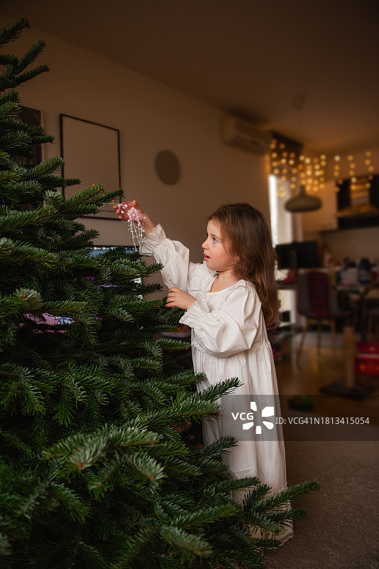 小女孩穿着复古的白色睡衣，把粉红色水母形状的玻璃玩具挂在真正的圣诞树上图片素材