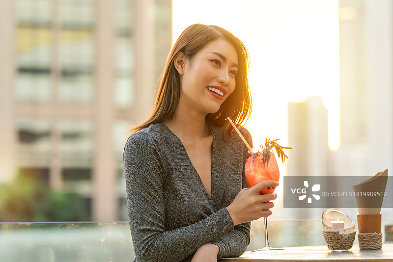 在温暖的晚霞中，一个亚洲女人在屋顶酒吧喝鸡尾酒图片素材