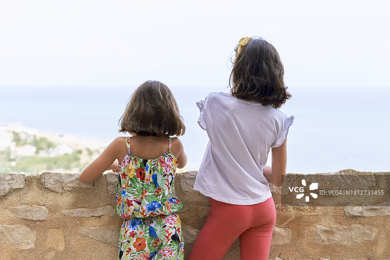 两个穿着便服的女孩站在石墙上，望向德尼亚城的后景。图片素材