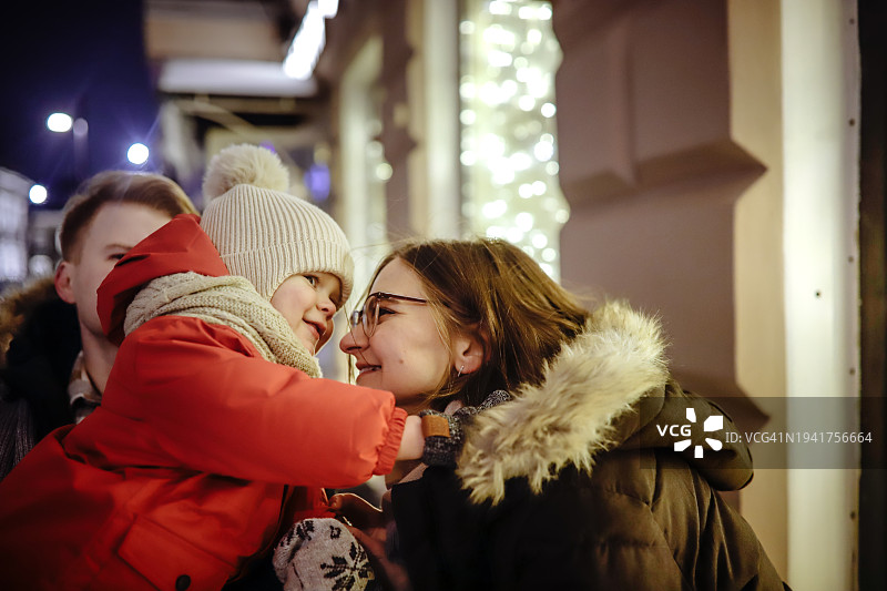 冬夜欢乐:快乐的年轻家庭，穿着舒适衣服的幼儿，探索灯火通明的街景图片素材