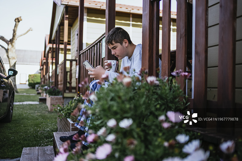 一个穿着睡衣的少年，一大早坐在满是鲜花的小木屋门廊的台阶上，一边喝着咖啡，一边玩着手机。图片素材