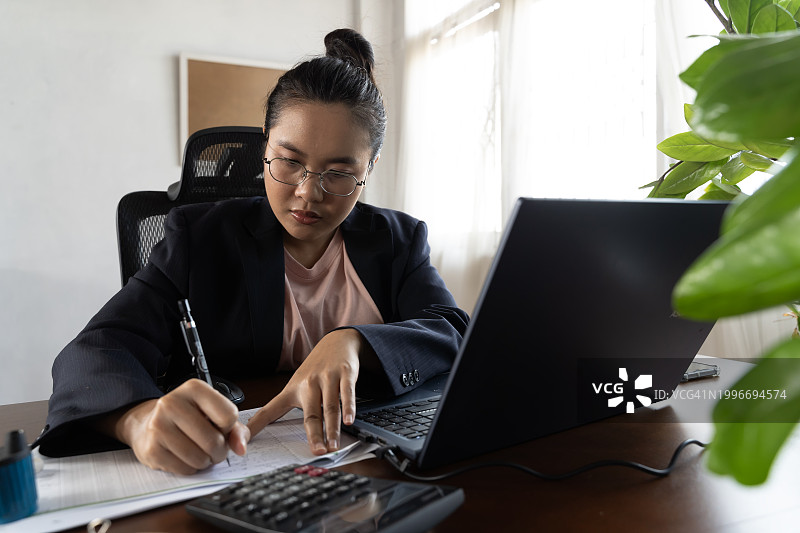 女商人坐在办公室的办公桌前，穿着西装，正在用笔记本电脑工作。她正在写文件。图片素材