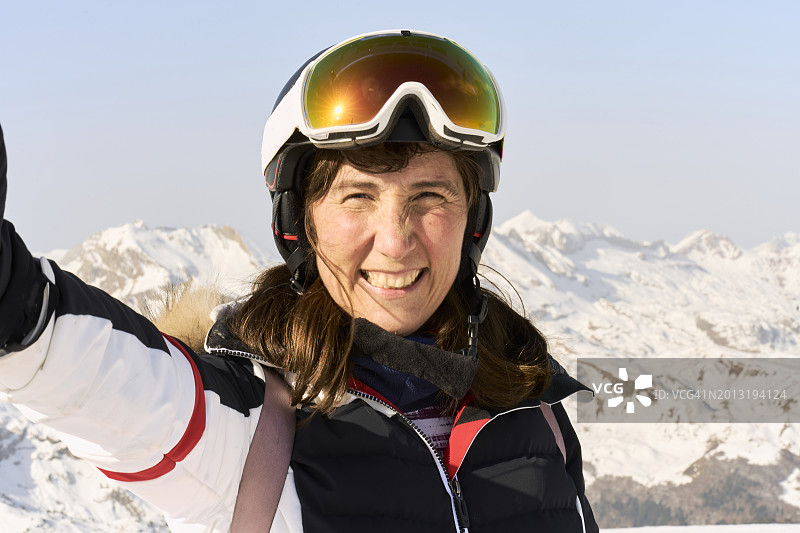 一个微笑的女人在山上的自拍特写，戴着滑雪装备，头盔和眼镜，看着相机，正面视图图片素材