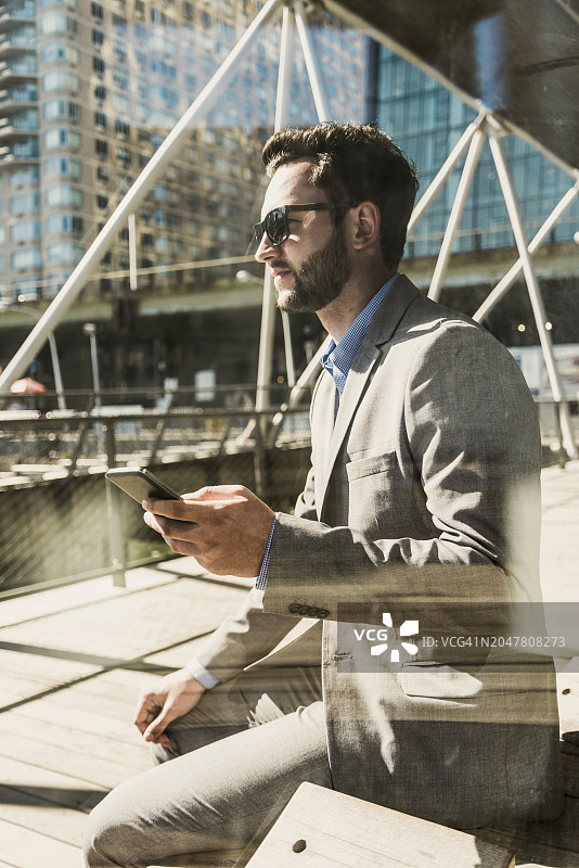 在阳光明媚的日子里，商人拿着智能手机坐在城市的长凳上图片素材
