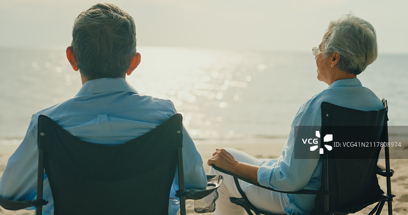 一对快乐的亚洲老夫妇坐在沙滩椅上欣赏海景，进行了深入的交谈。一个快乐的老年人享受退休后的旅行生活方式。图片素材